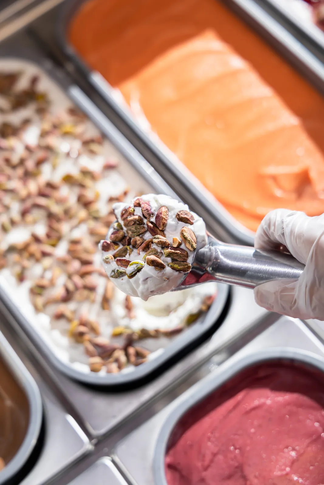 A scoop of pistachio-topped cream gelato lifted above colorful gelato pans